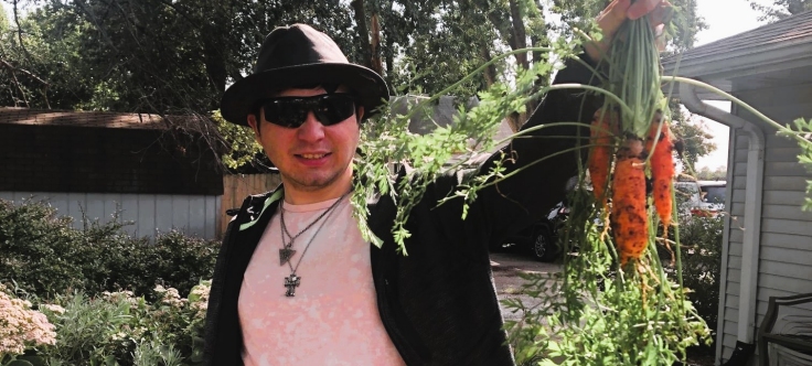 a young man proudly holds a bunch of carrots he grew in a garden